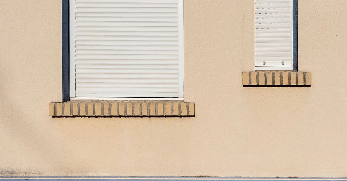 Una fachada de edificio beige con persianas blancas cerradas y un diseño arquitectónico minimalista.