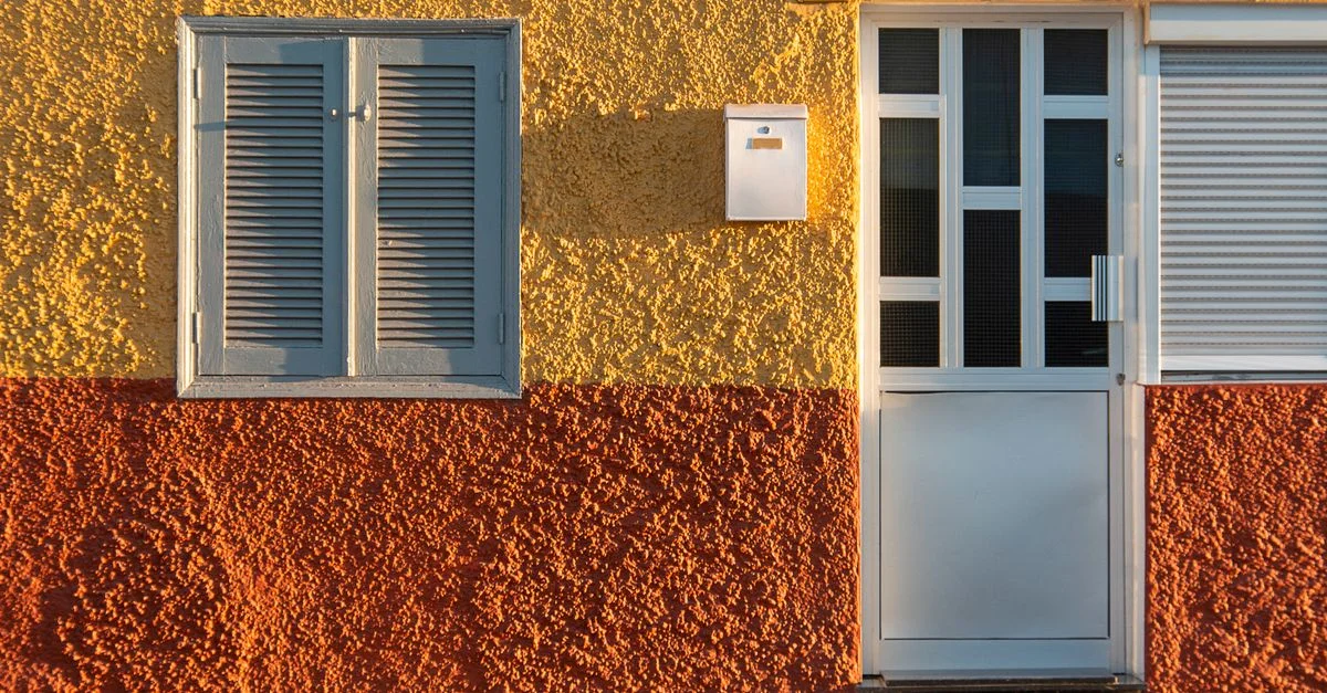 Fachada texturizada vibrante en amarillo y naranja con una persiana azul y una puerta blanca.