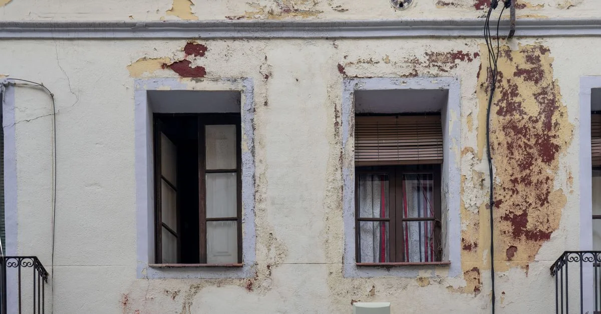 Primer plano de una fachada de edificio envejecida con pintura descascarada y tres ventanas.