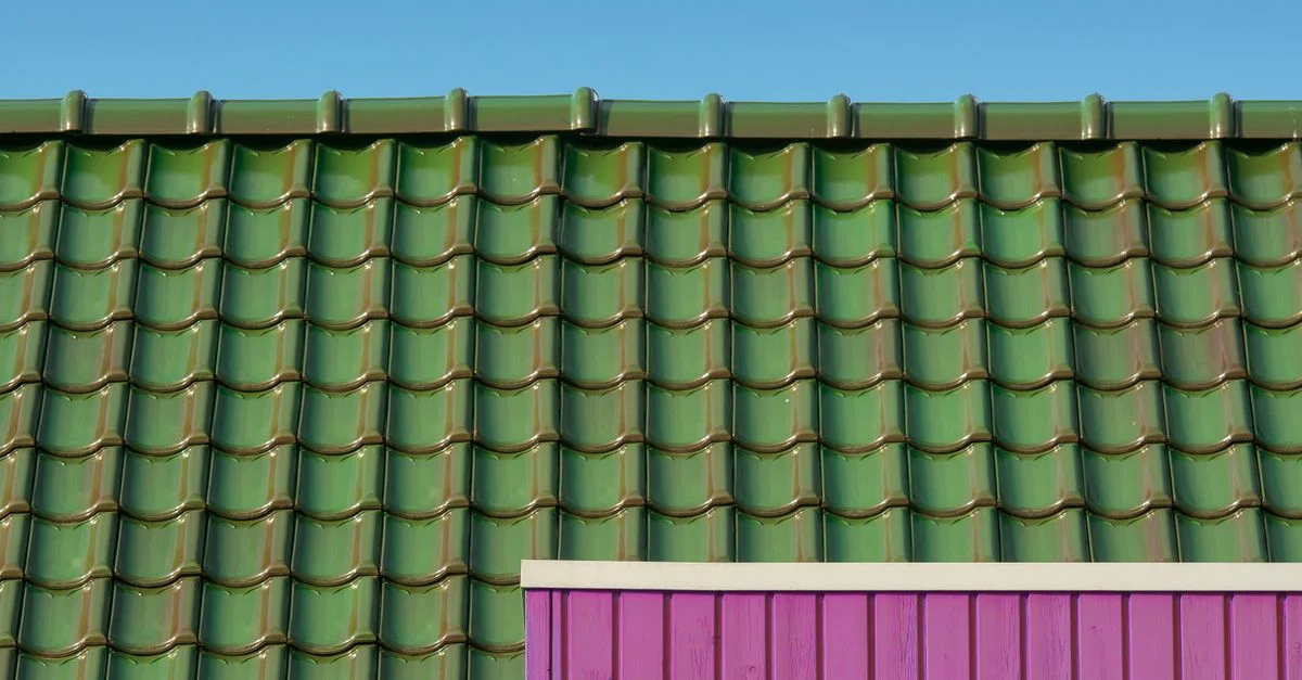 Un primer plano de un tejado verde de azulejos contra un cielo azul con una pared rosa contrastante.